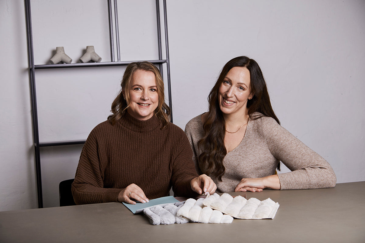 Zwei Frauen (links: Miriam, rechts: Ramona) sitzen an einem Tisch mit Stoffproben. Sie scheinen in eine Unterhaltung vertieft zu sein.