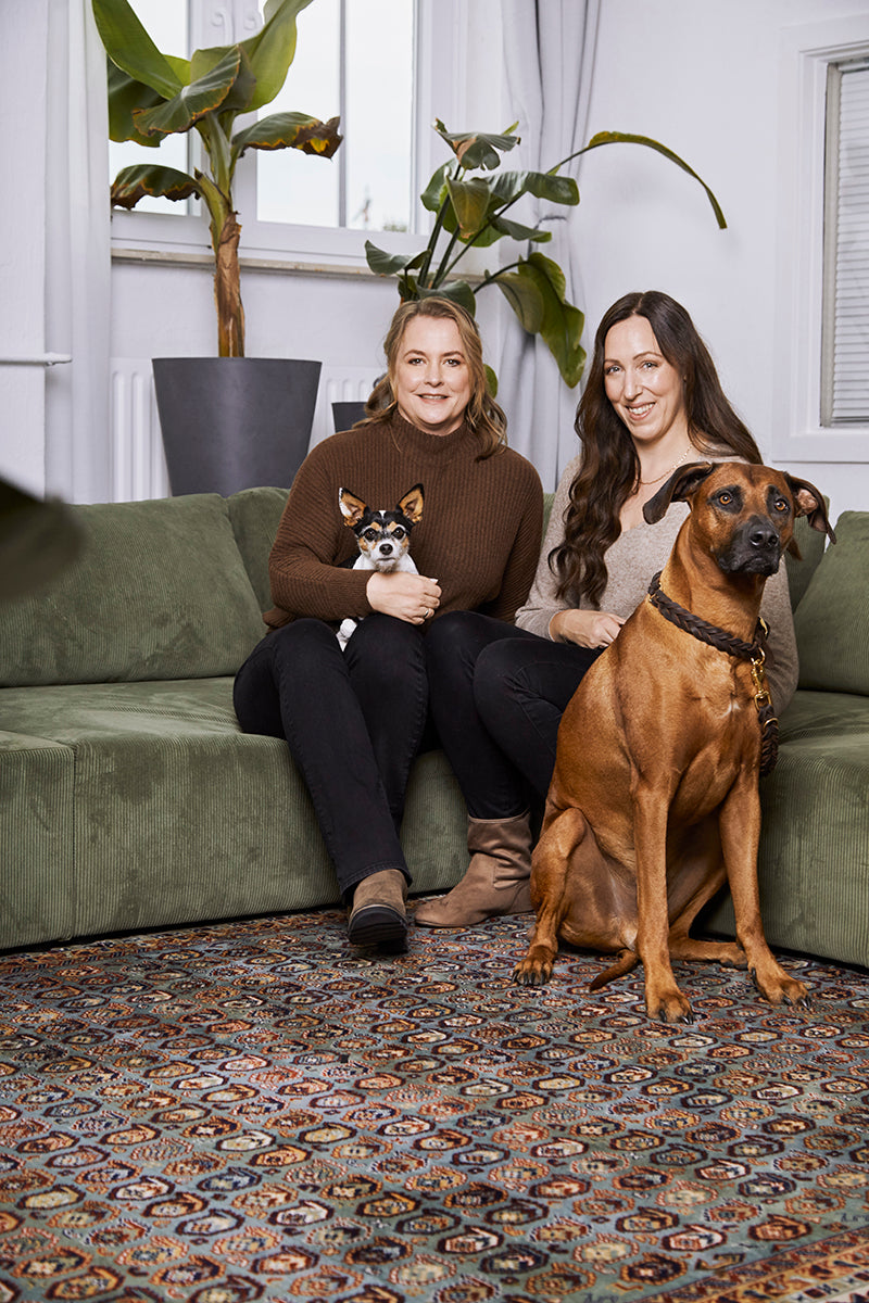 Zwei Frauen (links: Miriam, rechts: Ramona) sitzen mit ihren beiden Hunden zusammen auf einem Sofa.